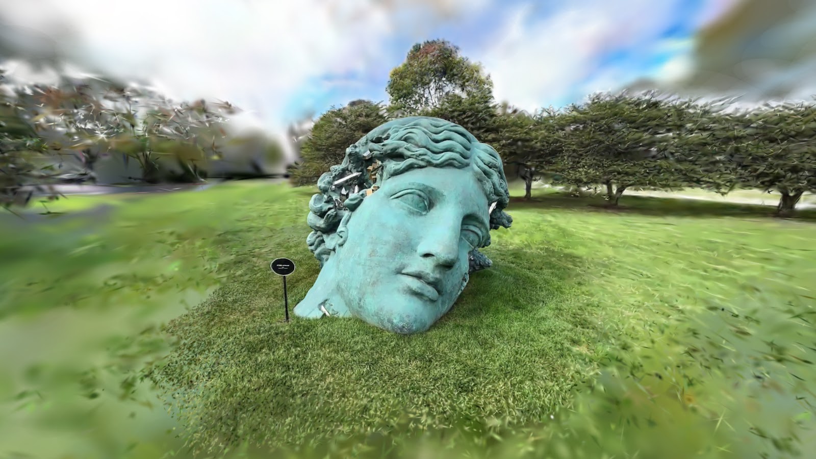 Gaussian splat capture of a giant bronze head sculpture on grass, with the typical fuzzy edges of a 3DGS reconstruction
